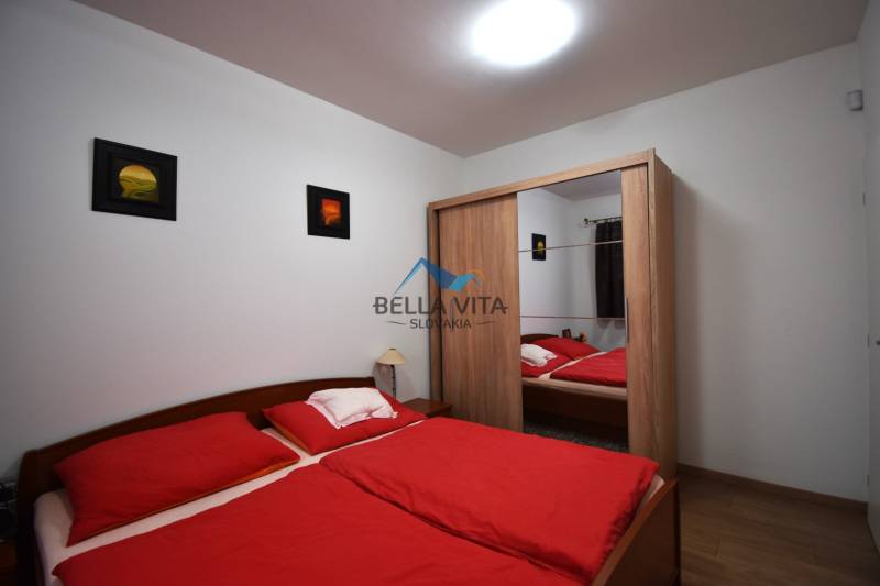 Spálňa s dvojposteľou, červeným povlečením, zrkadlovou skriňou a podlahou s dreveným dekorom.