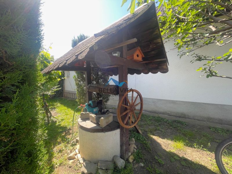 Studňa v záhrade pri rodinnom dome s drevenou strechou a dekoračným kolesom.