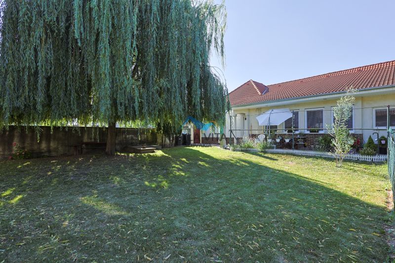 Rodinný dom s červenou strechou obklopený upravenou záhradou a veľkým stromom.